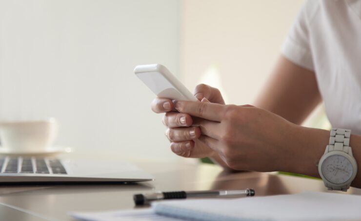 Close up image of modern cellphone in womans hand at desk with laptop. Office worker boring at workplace and playing games, writing in social networks. Businesswoman looking important contact on phone